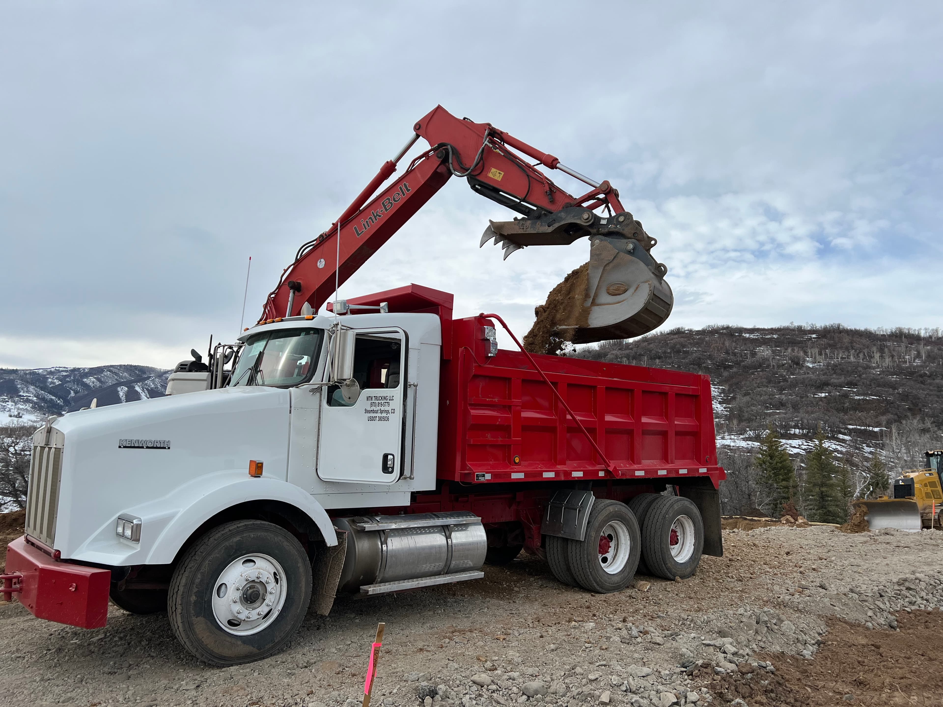 An MTM dump truck loading aggregate with its red grapple arm against a snow-dusted Colorado ridgeline.