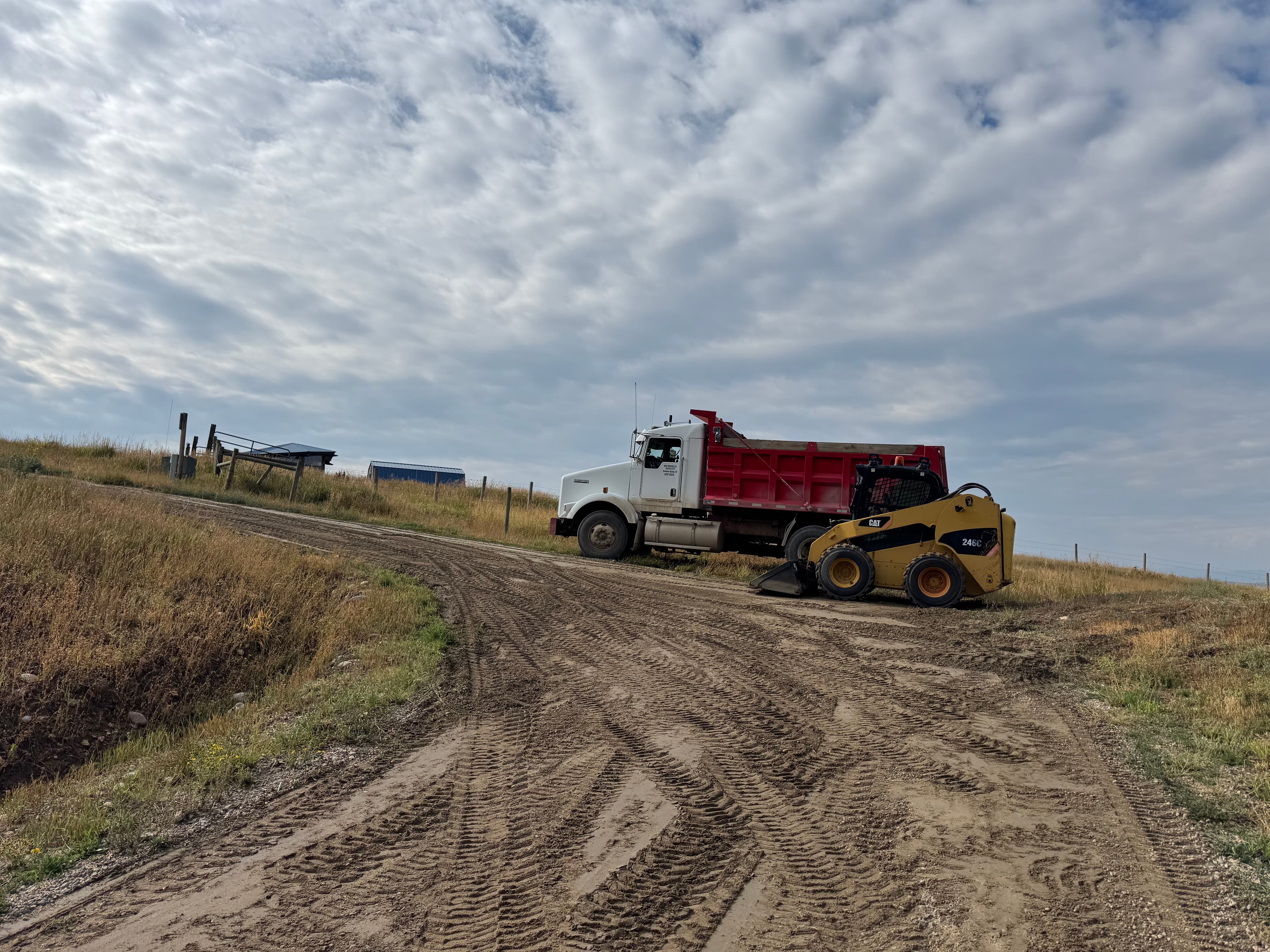 A red-and-white truck and a yellow skid steer working a wide Colorado prairie site under a big cloud sky.